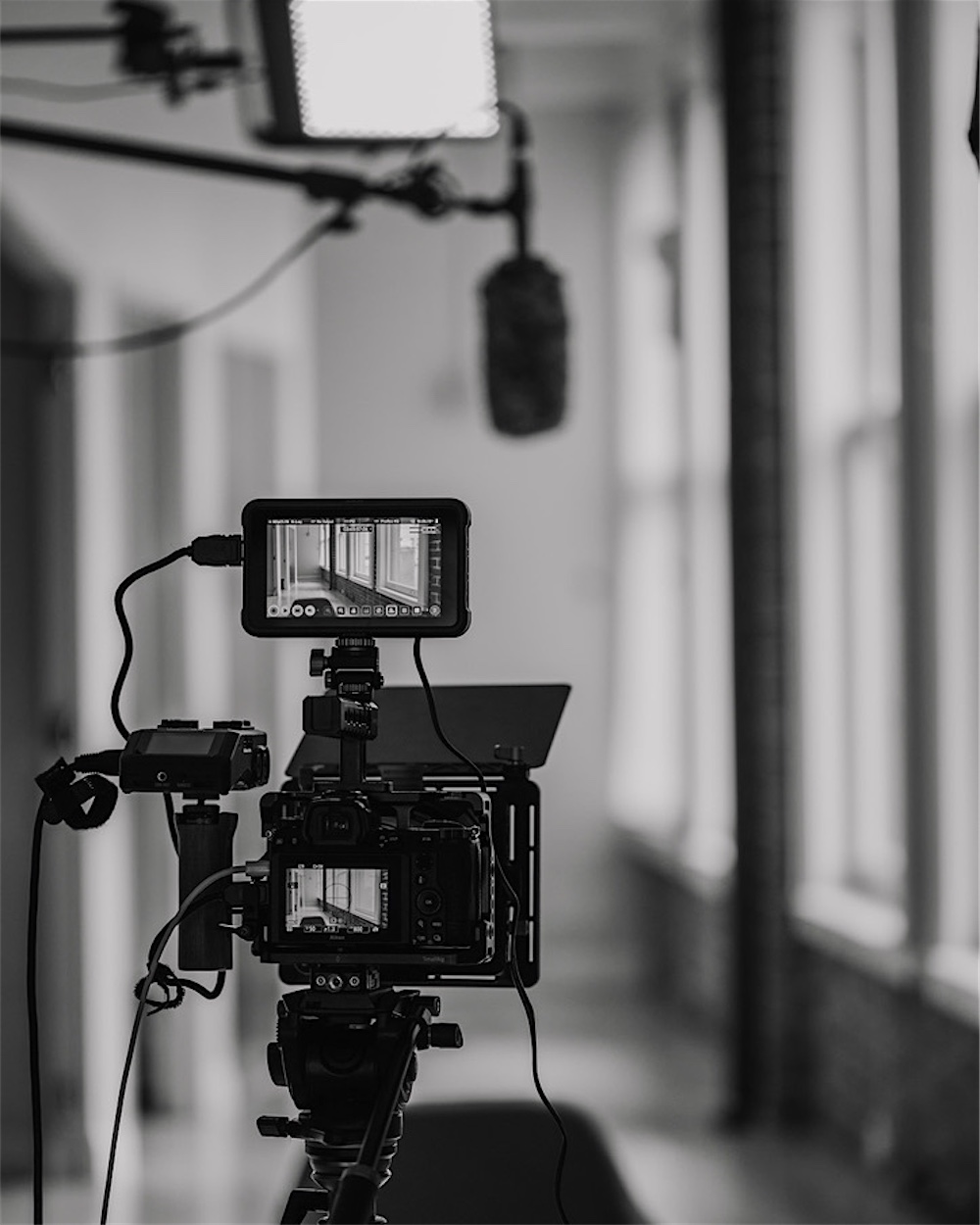 A cinema camera rig with a monitor, light, and microphone set up in a bright corridor, in black and white