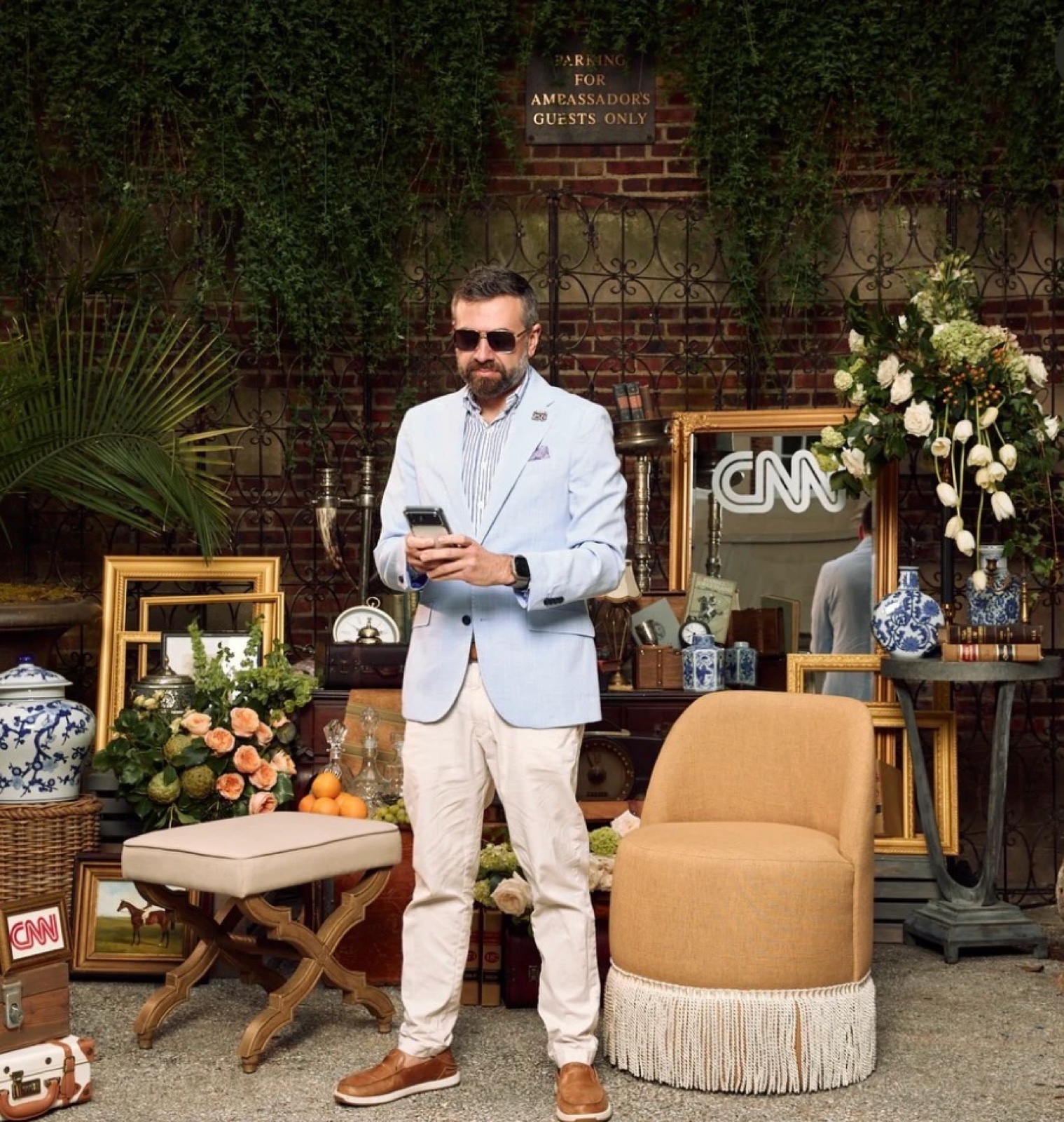 Matt Shupe in a light blue blazer checking his phone in a courtyard staged with antiques, flowers and CNN signage