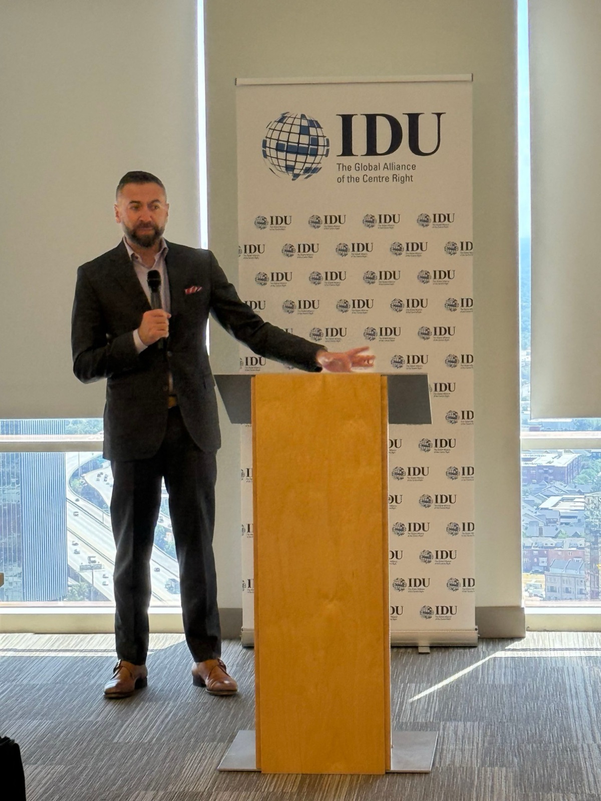 Matt Shupe speaking beside a lectern in front of an International Democrat Union banner, a city skyline behind the glass