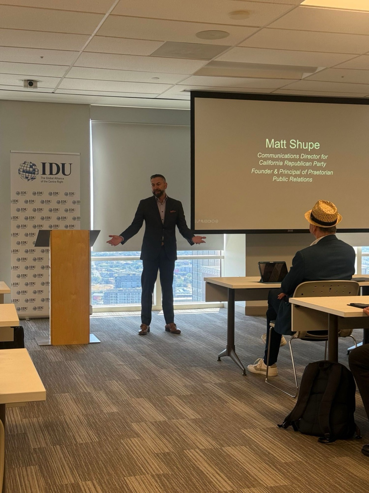 Matt Shupe addressing a seminar room at an International Democrat Union session, his name and titles on the screen